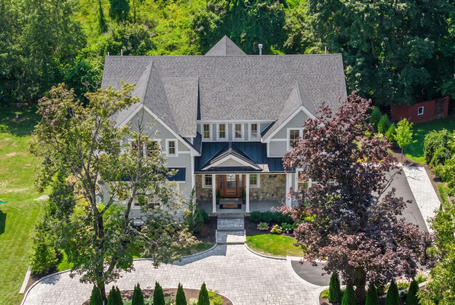 Aerial close-up of the front elevation showing the roofline, board-and-batten entry gable, and circular paver drive framed by mature summer trees