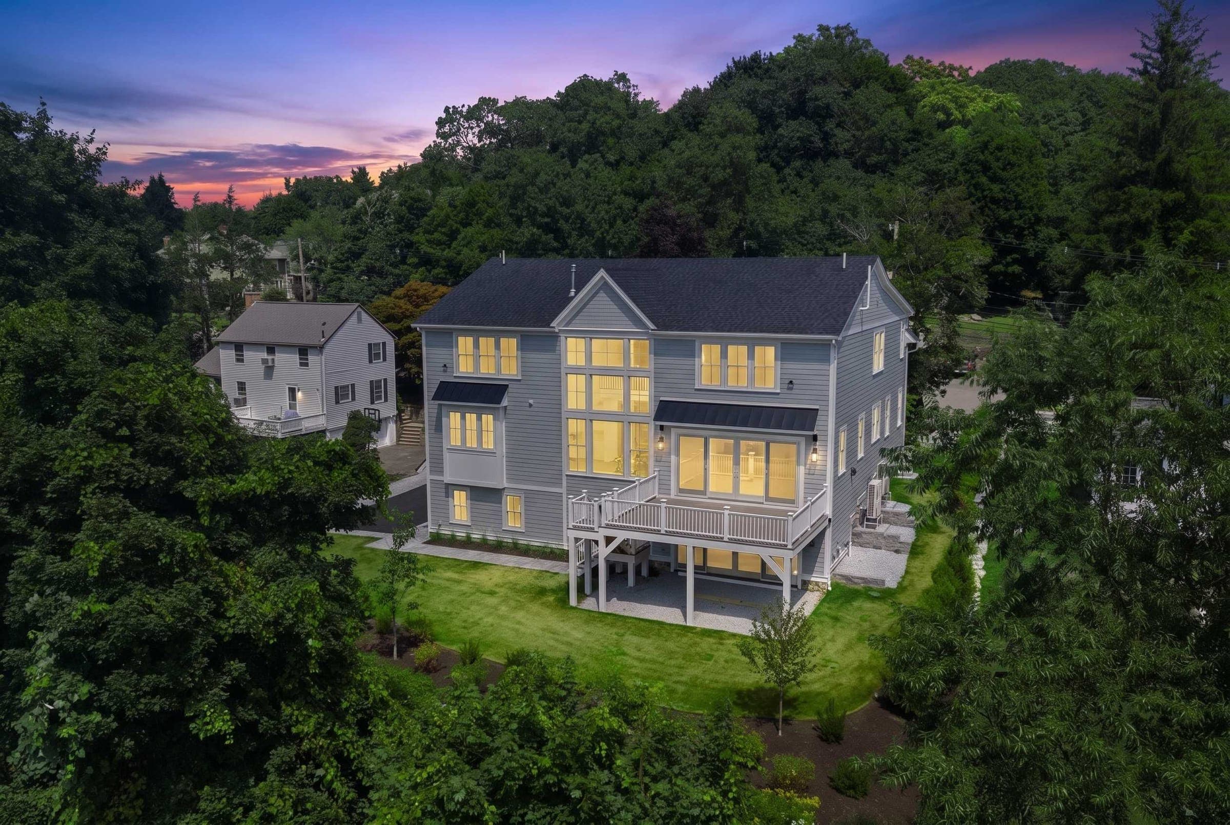 320 Concord Avenue at dusk — the home glowing warm against a sunset sky, nestled privately within mature summer woods